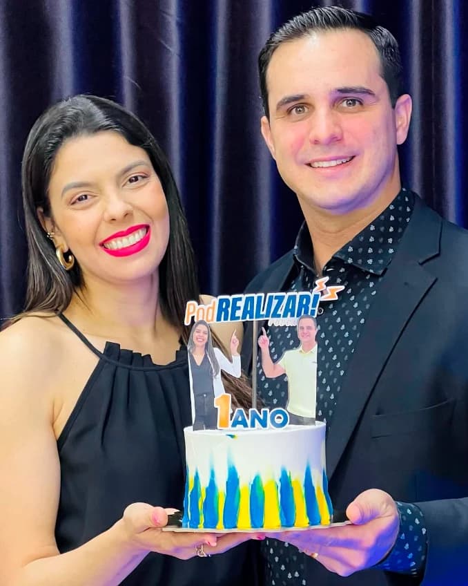 Karina Esteves ao lado do co-apresentador segurando o bolo de comemoração de 1 ano do PodRealizar.